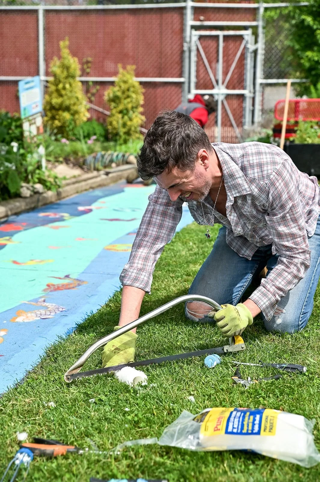 Adam Lekach with gloves kneels on the grass, smiling, using a hand saw on a piece of wood outdoors with colorful painted fish on a blue background, in a garden with a fence and bushes.