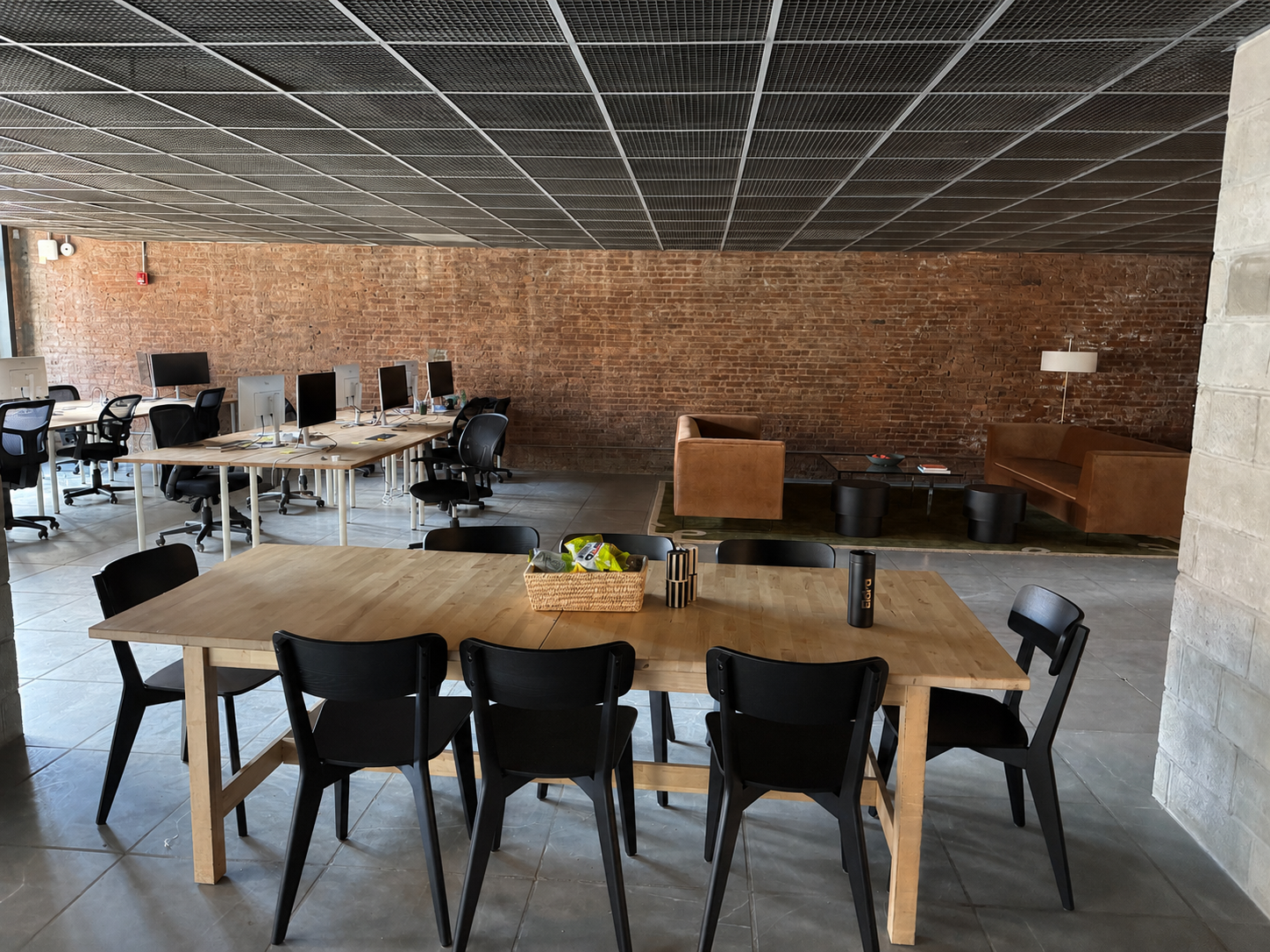 An office space with a wooden table and black chairs in the foreground, a seating area with a leather sofa and armchair, multiple workstations with monitors, and a brick accent wall, all under a dark grid ceiling.
