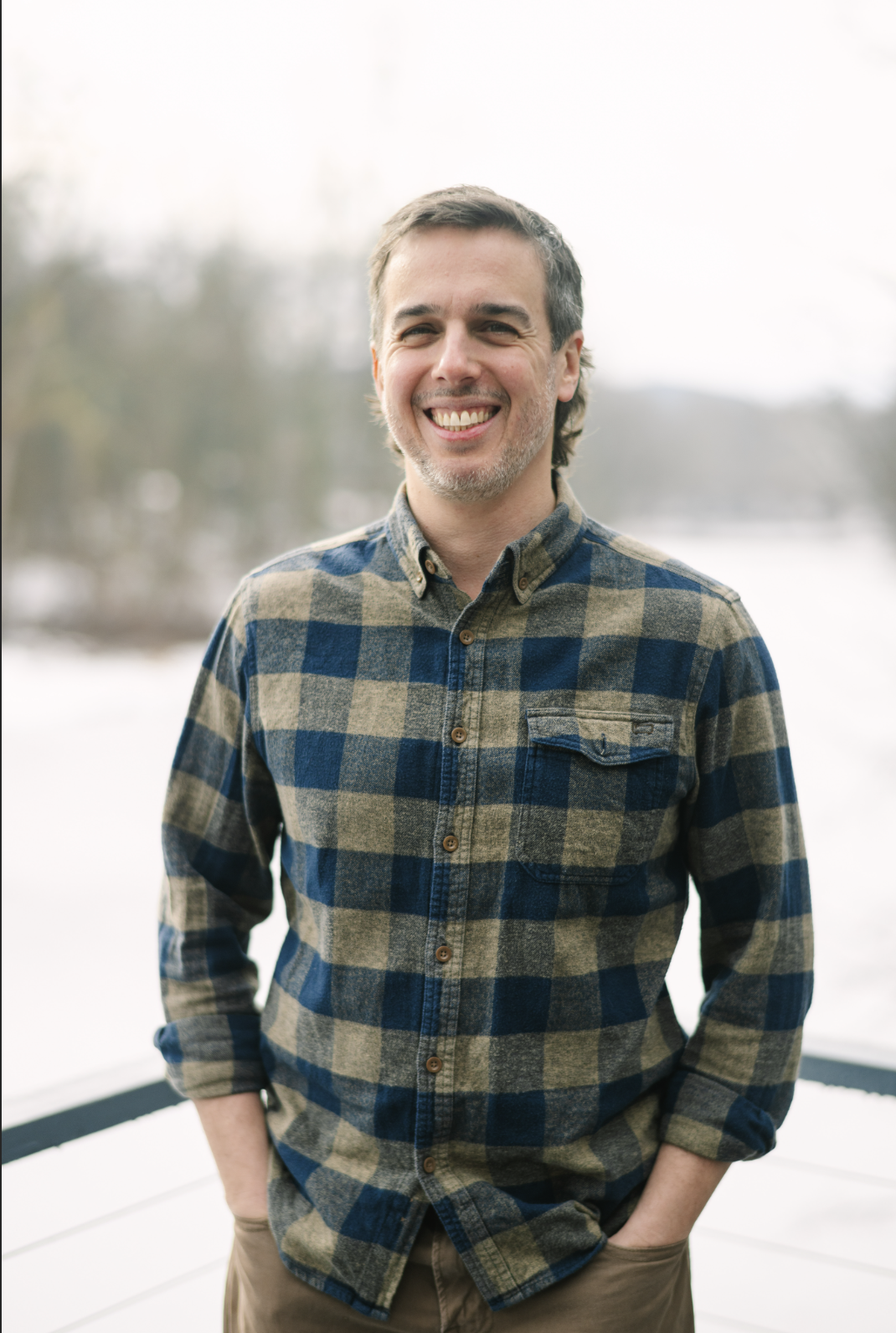 Adam Lekach in front of a body of water and trees, wearing a blue and beige plaid shirt with rolled-up sleeves.
