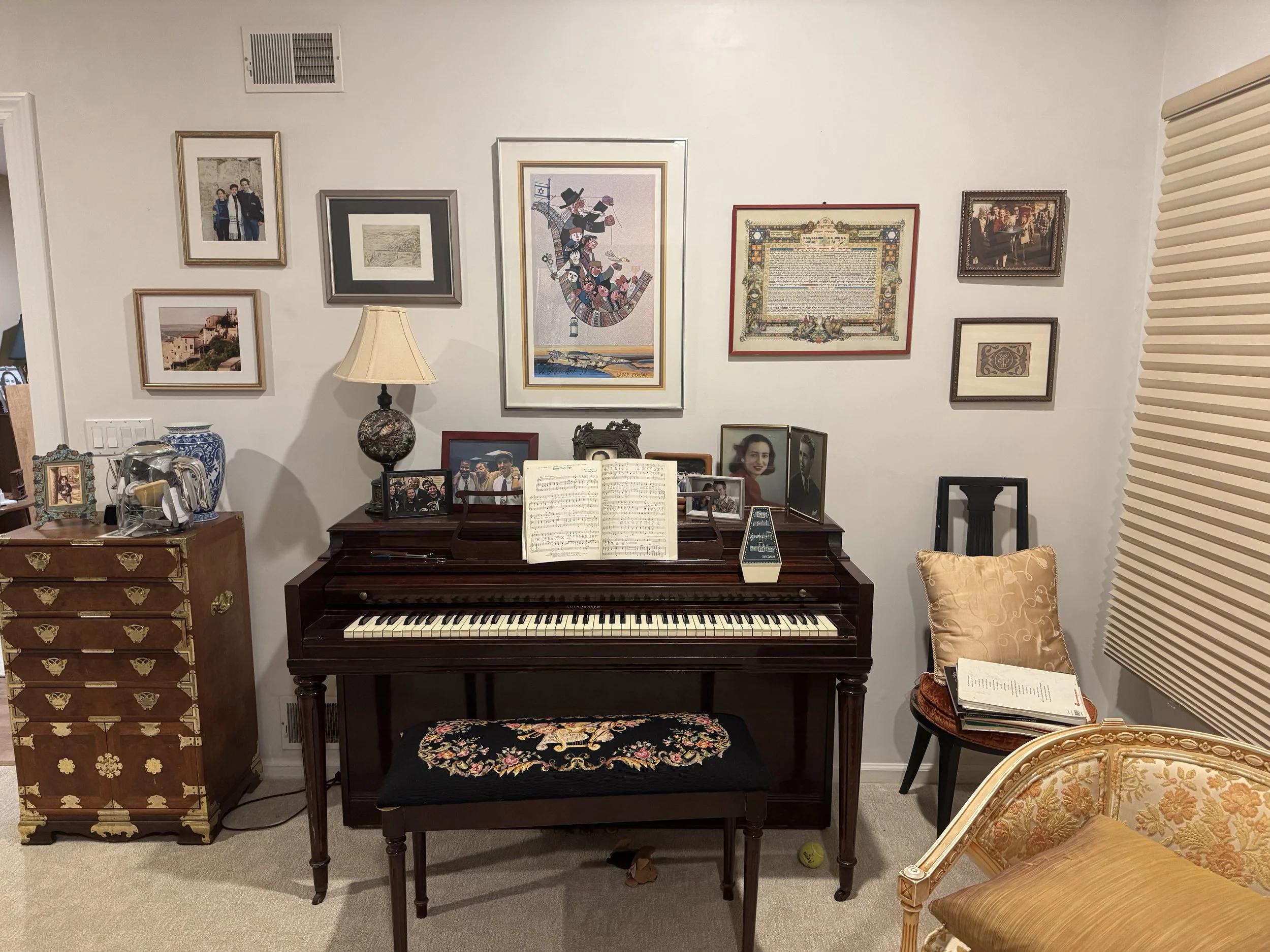 A living room wall with multiple framed photos and artwork, a piano with music sheets, a table with framed photos, and a decorative chair and lamp.