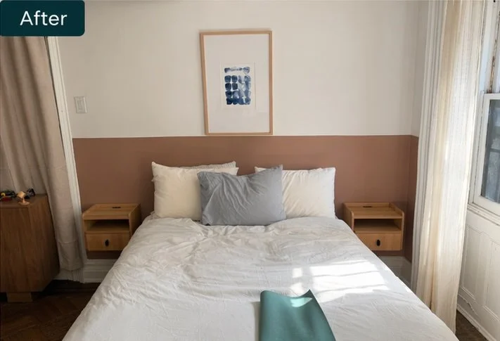 A neatly made bed with white and gray pillows against a two-tone beige and white wall, with a framed abstract art piece above it. There are two small wooden nightstands on either side, and sunlight streams through a window with light-colored curtains.