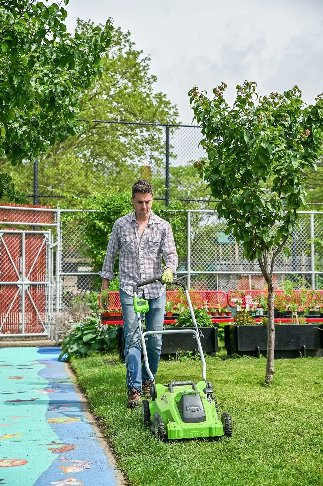 A man wearing a plaid shirt and jeans uses a green and black electric lawn mower to cut grass in a backyard with trees, a chain-link fence, and potted plants.