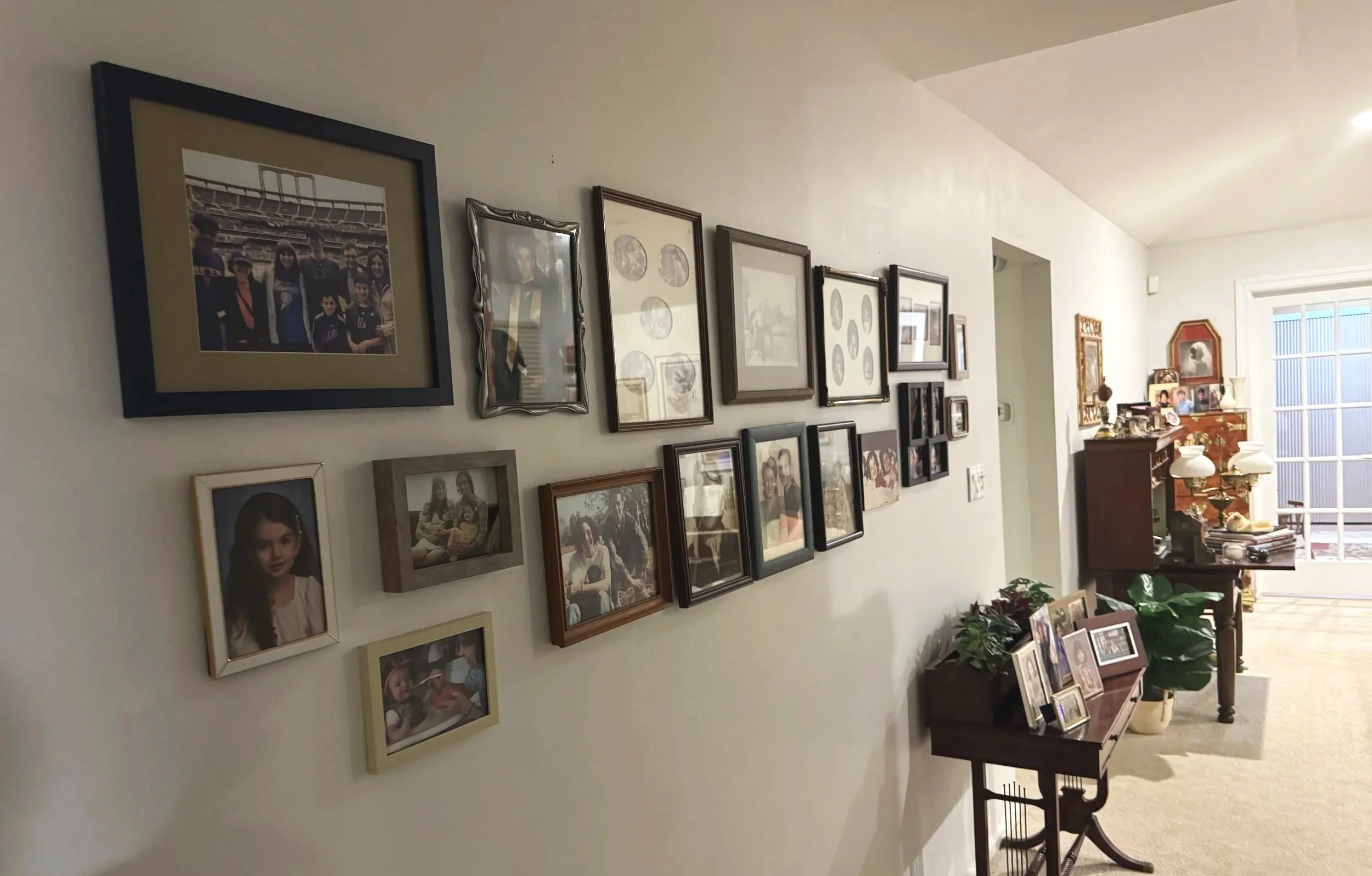 A wall decorated with numerous framed family photographs in a living room, with a corner of a wooden table visible. In the background, there's a large window with blinds, a sideboard filled with picture frames, lamps, and decorative items, and plants on the floor.