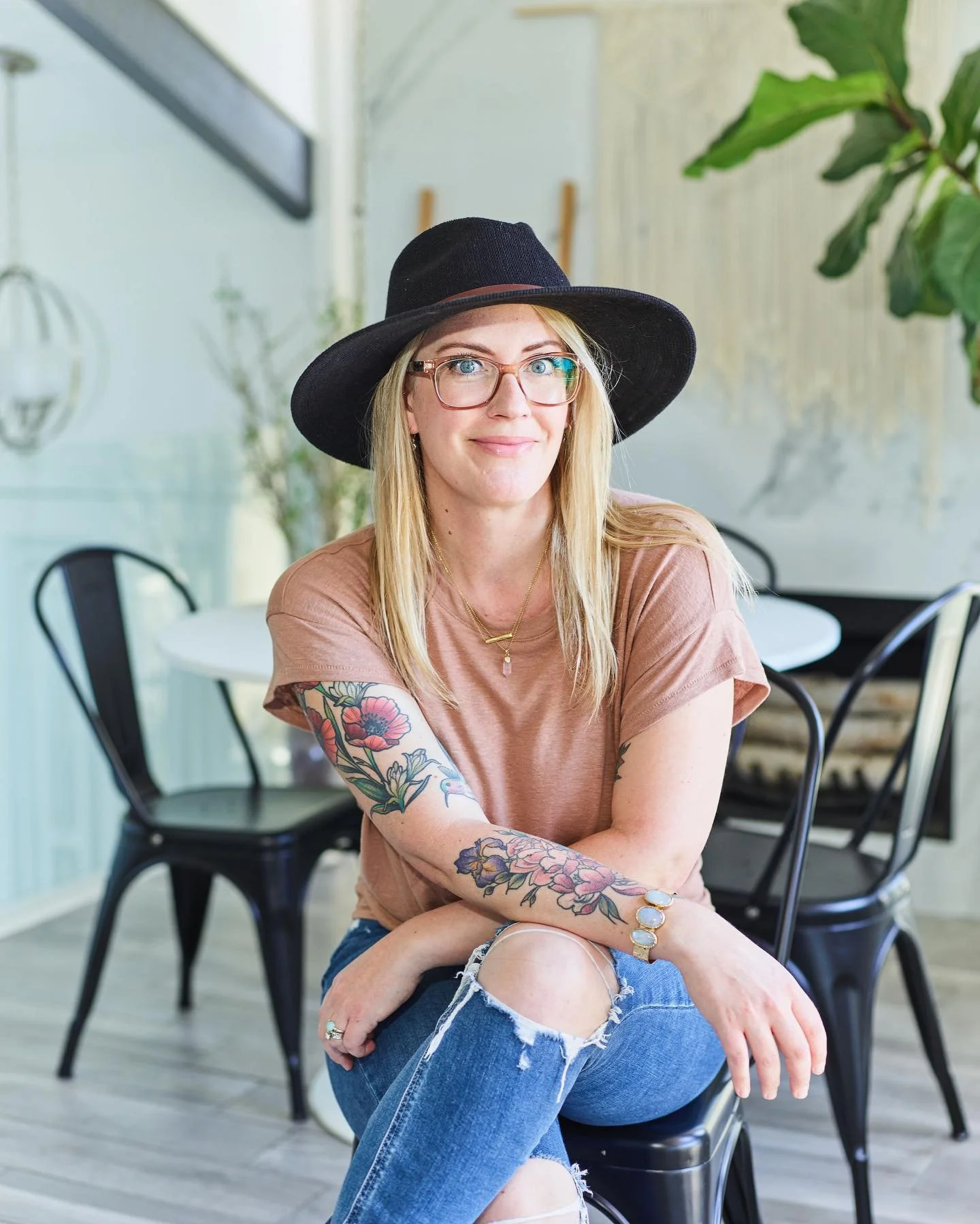 A woman with long blonde hair, wearing a black wide-brimmed hat, glasses, a pink t-shirt, and ripped jeans, sitting in a modern, light-filled room with plants and black chairs.