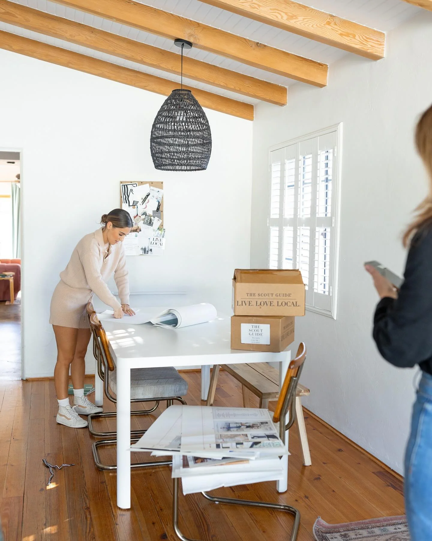 A woman in beige clothing bends over a table looking at a large rolled-up paper, in a room with wooden floors and white walls. There are brown cardboard boxes on the table labeled "The Scout Guide" and a bulletin board on the wall behind her. Another person, partially visible, stands on the right looking at a phone.