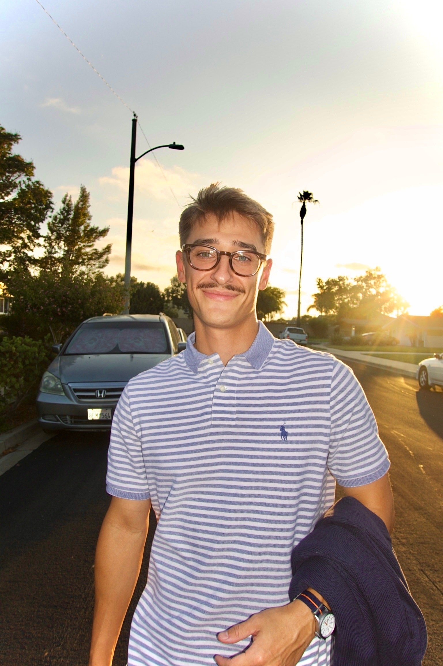A young man with glasses and a striped polo shirt smiling outside during sunset, with cars, trees, and streetlights in the background.