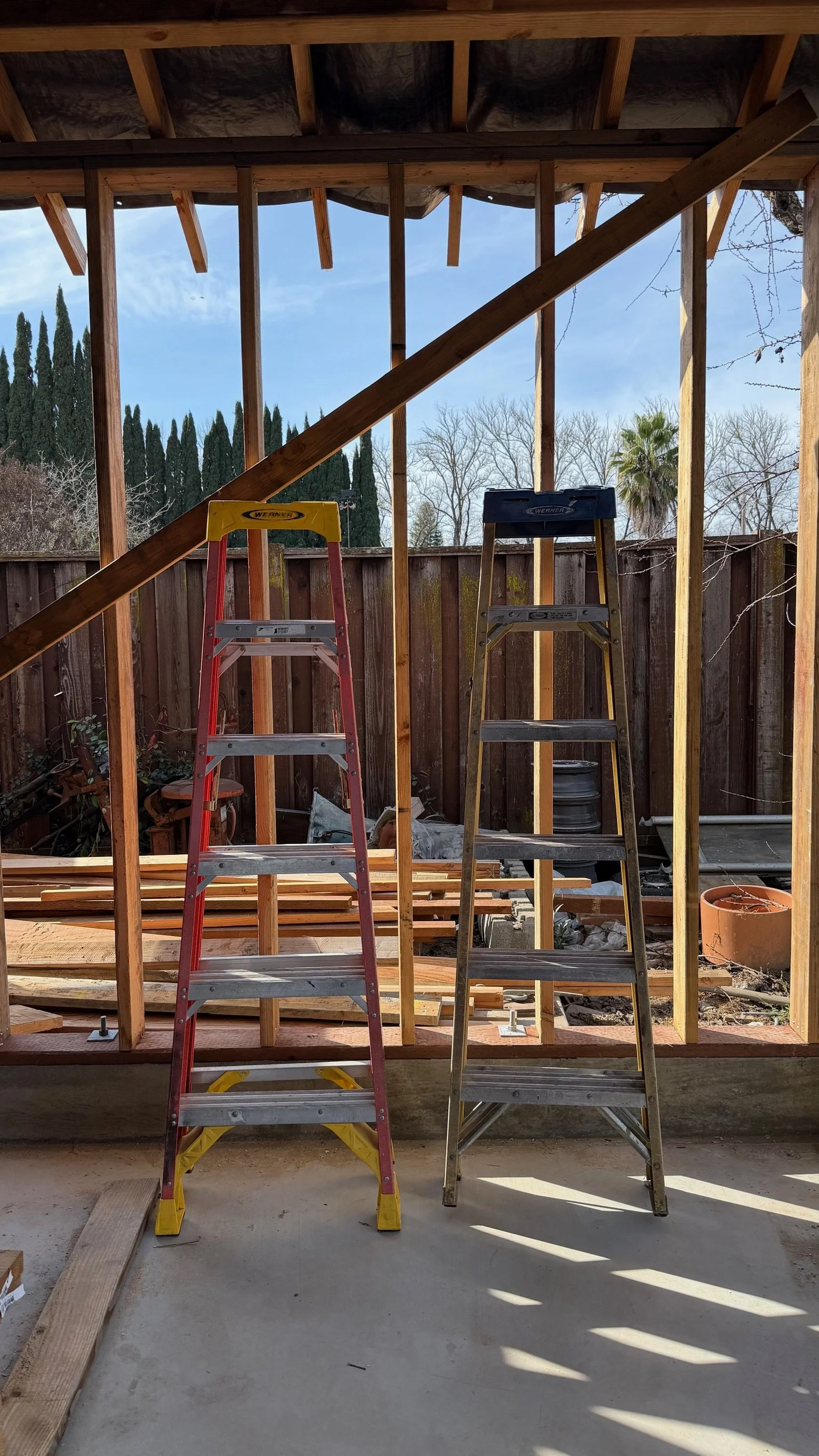 Framed view of a construction site with two ladders, one red and one silver, set inside a wooden frame for a building under construction. The background features a wooden fence and trees, with a clear sky.