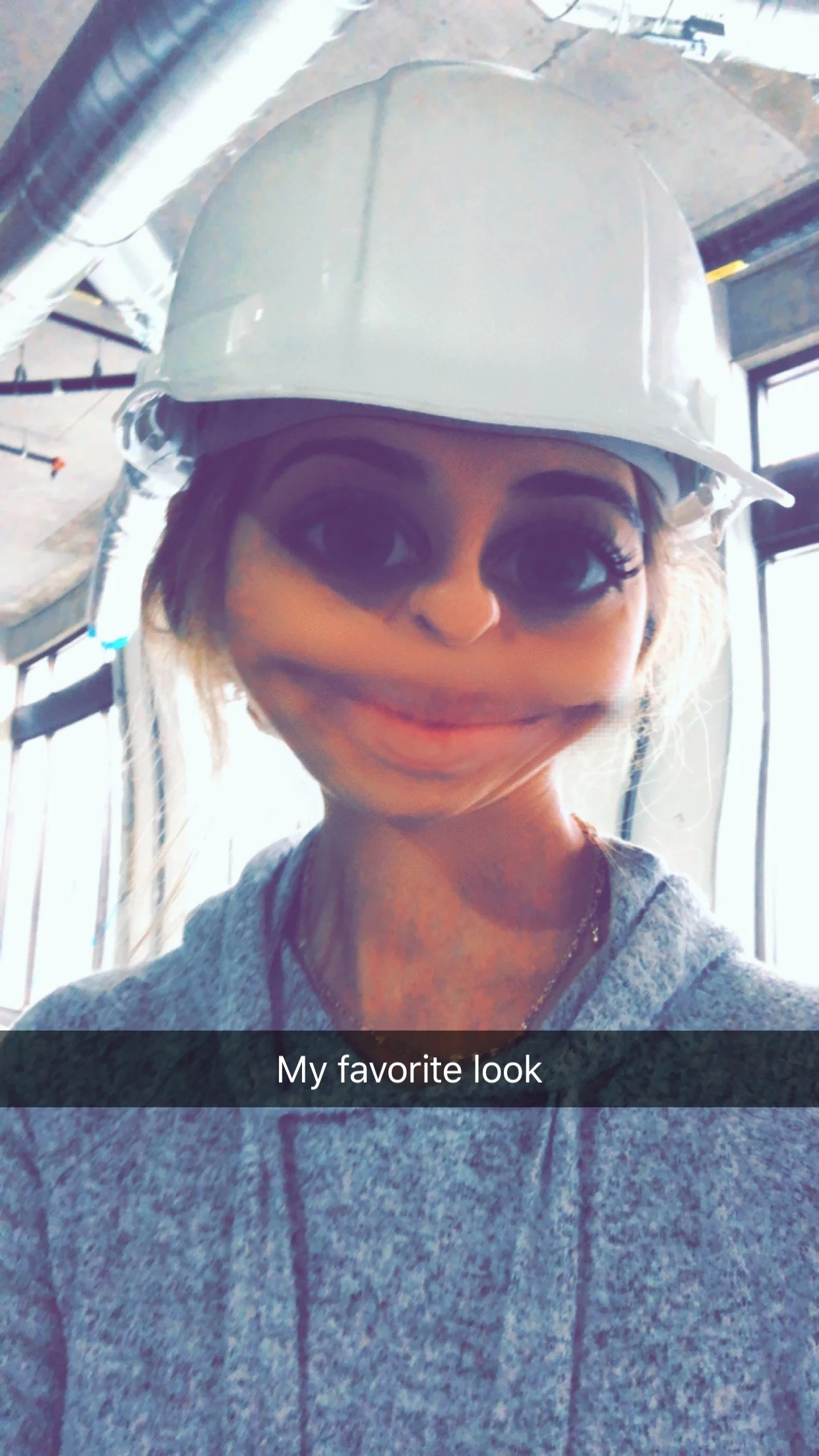 A woman wearing a white safety helmet and a gray hoodie, smiling with a filter that enlarges her eyes, with construction pipes and a building under construction in the background.