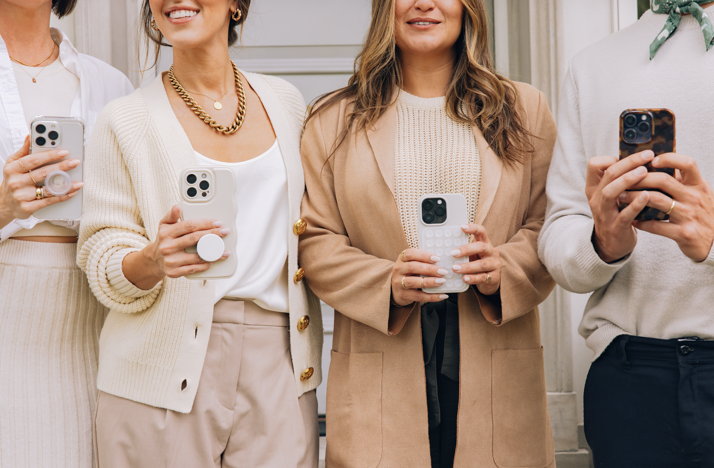 Group of women holding smartphones, dressed in neutral tones, smiling, and standing together outdoors.