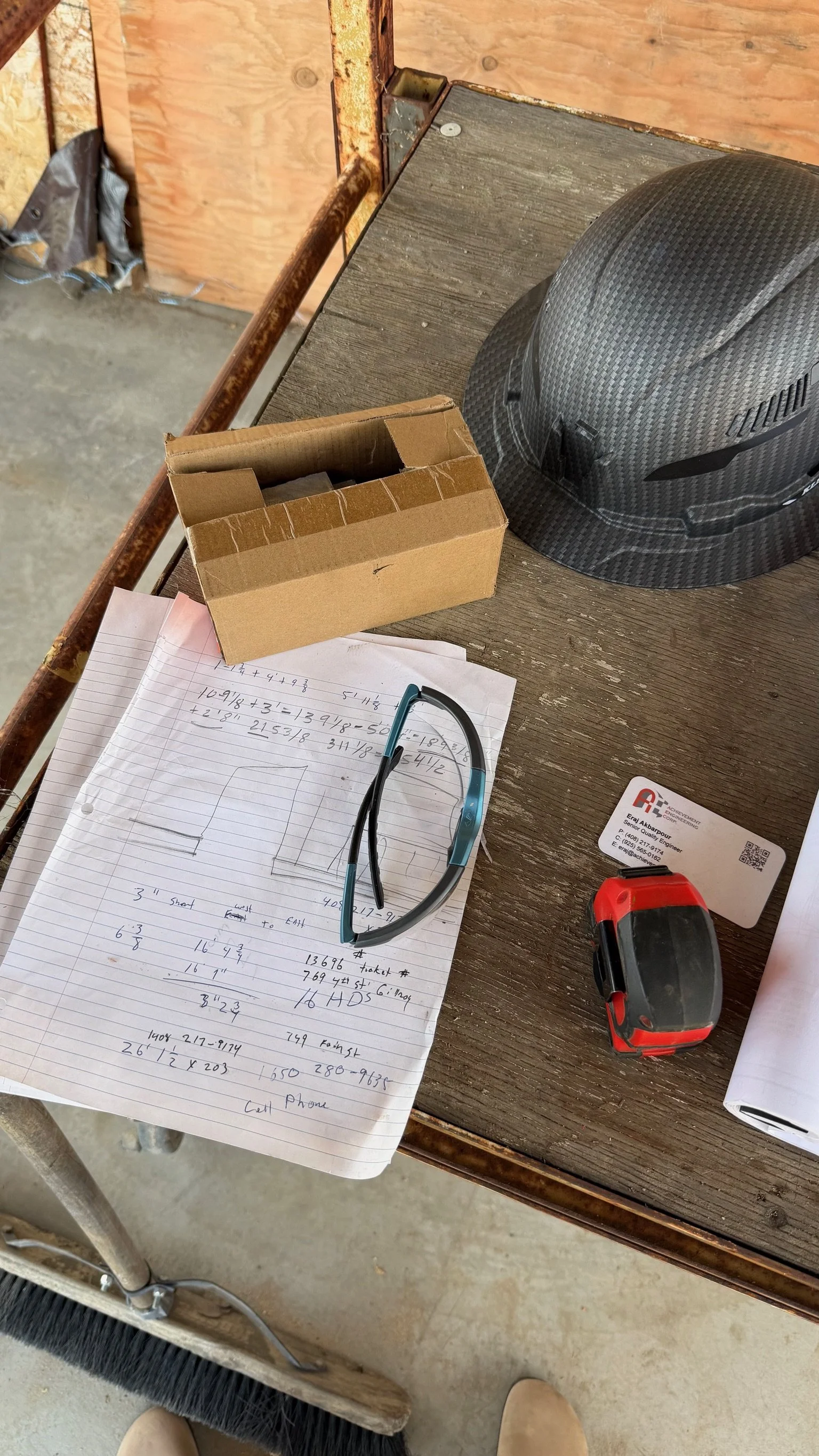 A cluttered workspace with a piece of lined paper containing handwritten notes and calculations, a pair of safety glasses, a red and black measuring tape, a business card, a cardboard box, a helmet with a carbon fiber pattern, and a few sheets of paper on a wooden surface, with a rough concrete floor underneath.