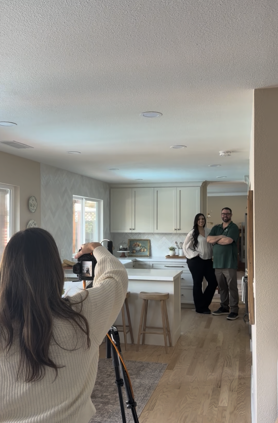 A woman is taking a photo of a smiling man and woman standing in a kitchen with light-colored cabinetry and a white island.