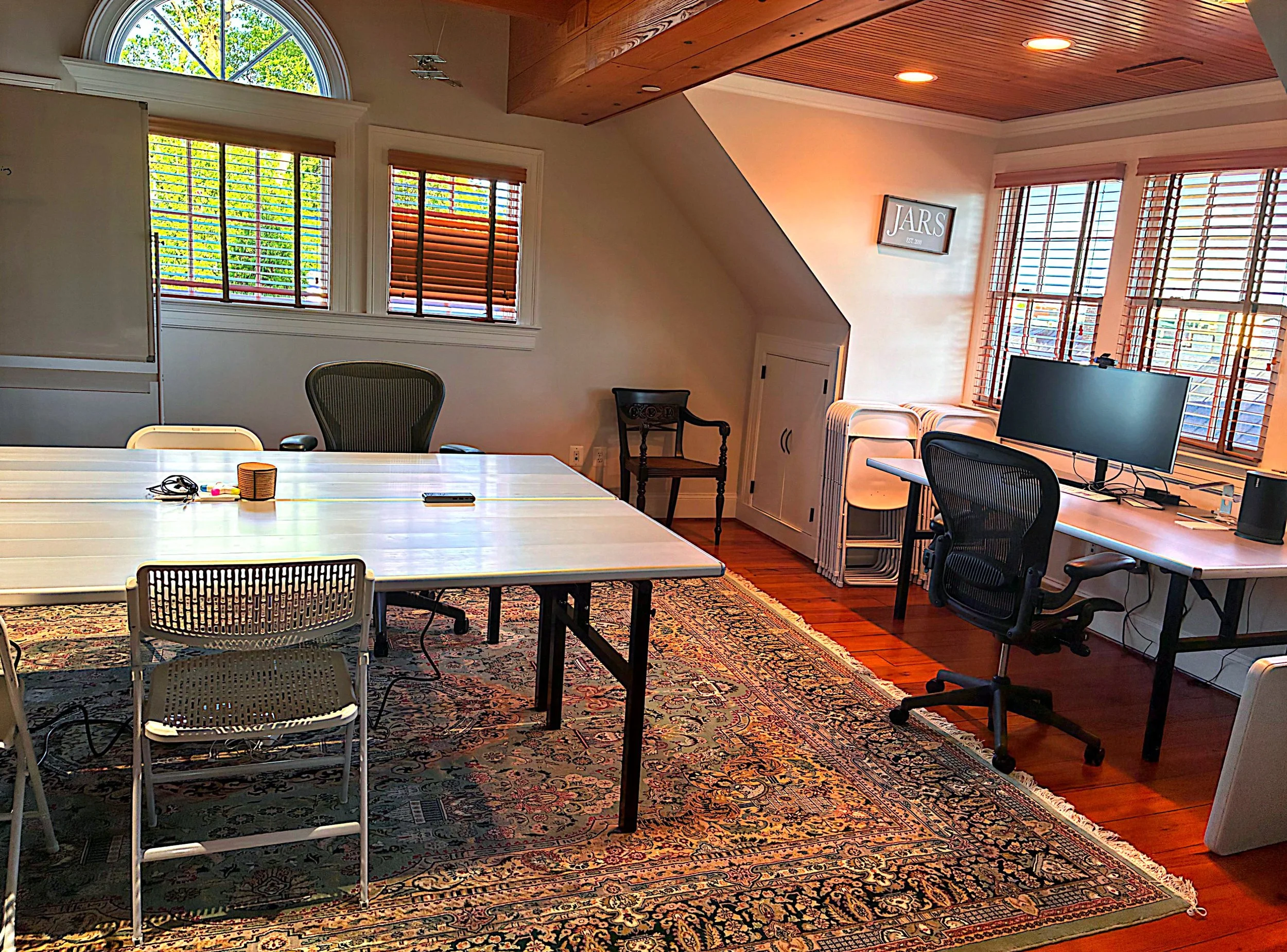 Office or conference room with wooden ceiling, multiple windows with blinds, a table with chairs, a beige carpet with traditional pattern, a monitor on a desk, and a framed sign on the wall reading "JARS EST. 2018."