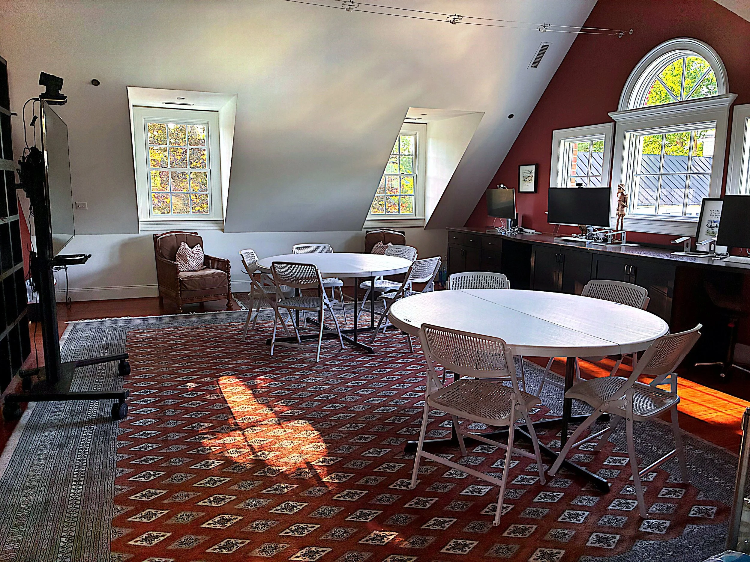 A bright room with a sloped ceiling and multiple windows letting in natural light. There are round tables and white chairs, a red patterned rug, and a black cabinet with electronic equipment. The windows show trees outside, and a red wall has framed pictures and monitors.