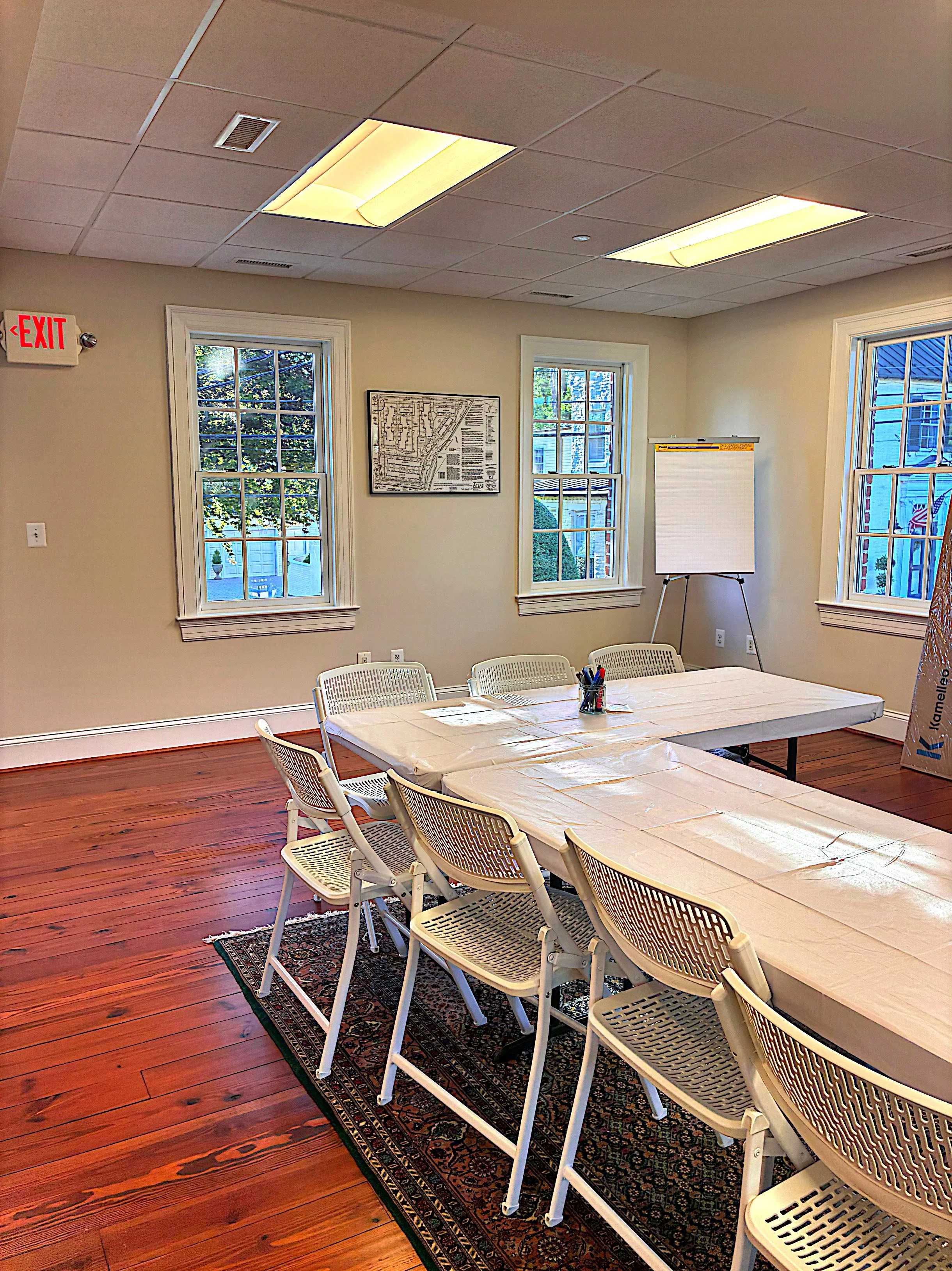 A meeting room with six white plastic chairs around a rectangular table covered with a plastic tablecloth. There is a black patterned rug under the table. The room has three windows with white trim, a framed map on the wall, a whiteboard on a stand, and a red exit sign on the wall. The ceiling has square panels with two fluorescent lights.