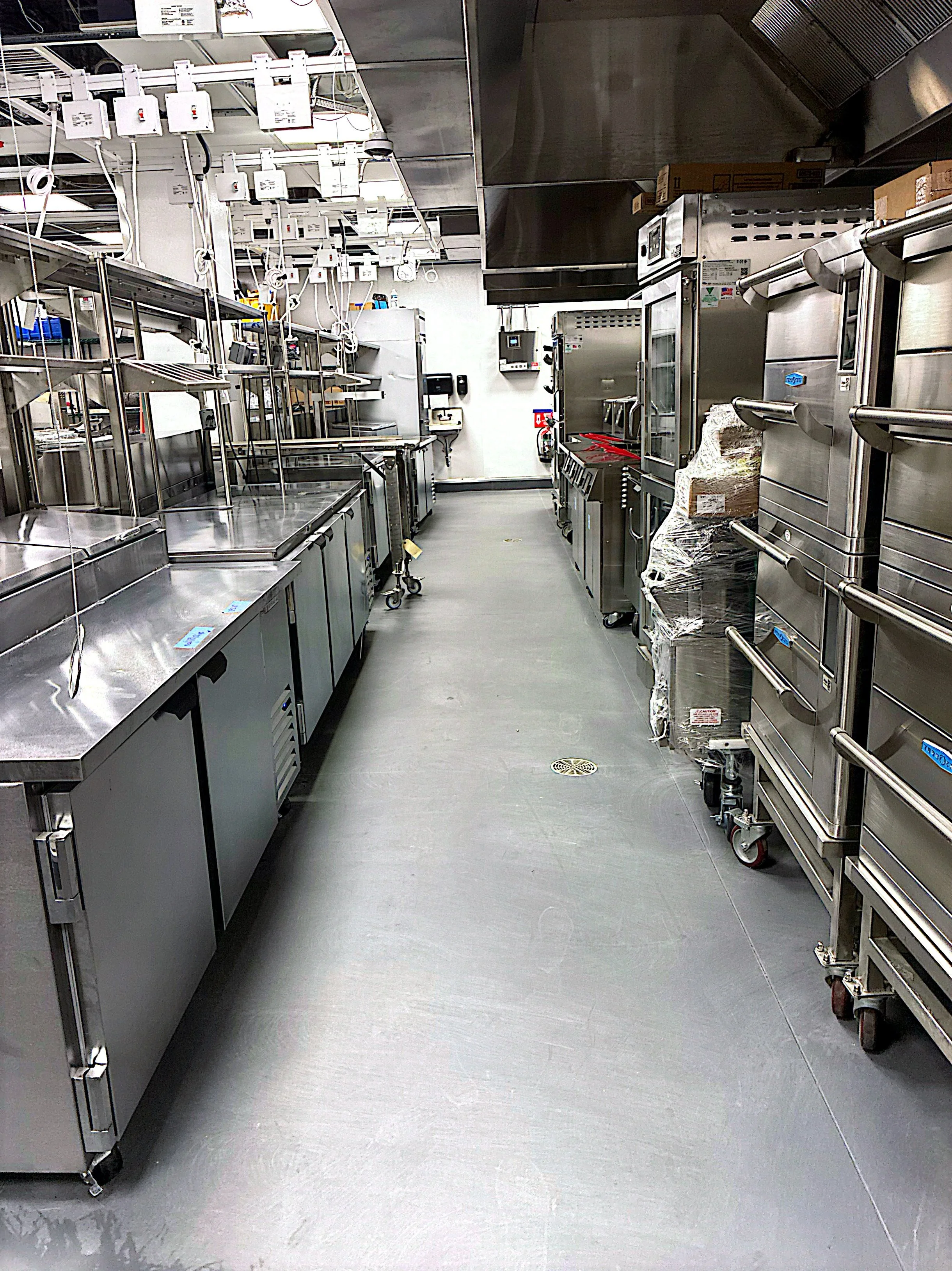 An empty commercial kitchen with stainless steel appliances and equipment, including ovens, shelves, and carts, arranged along the walls and across the room.