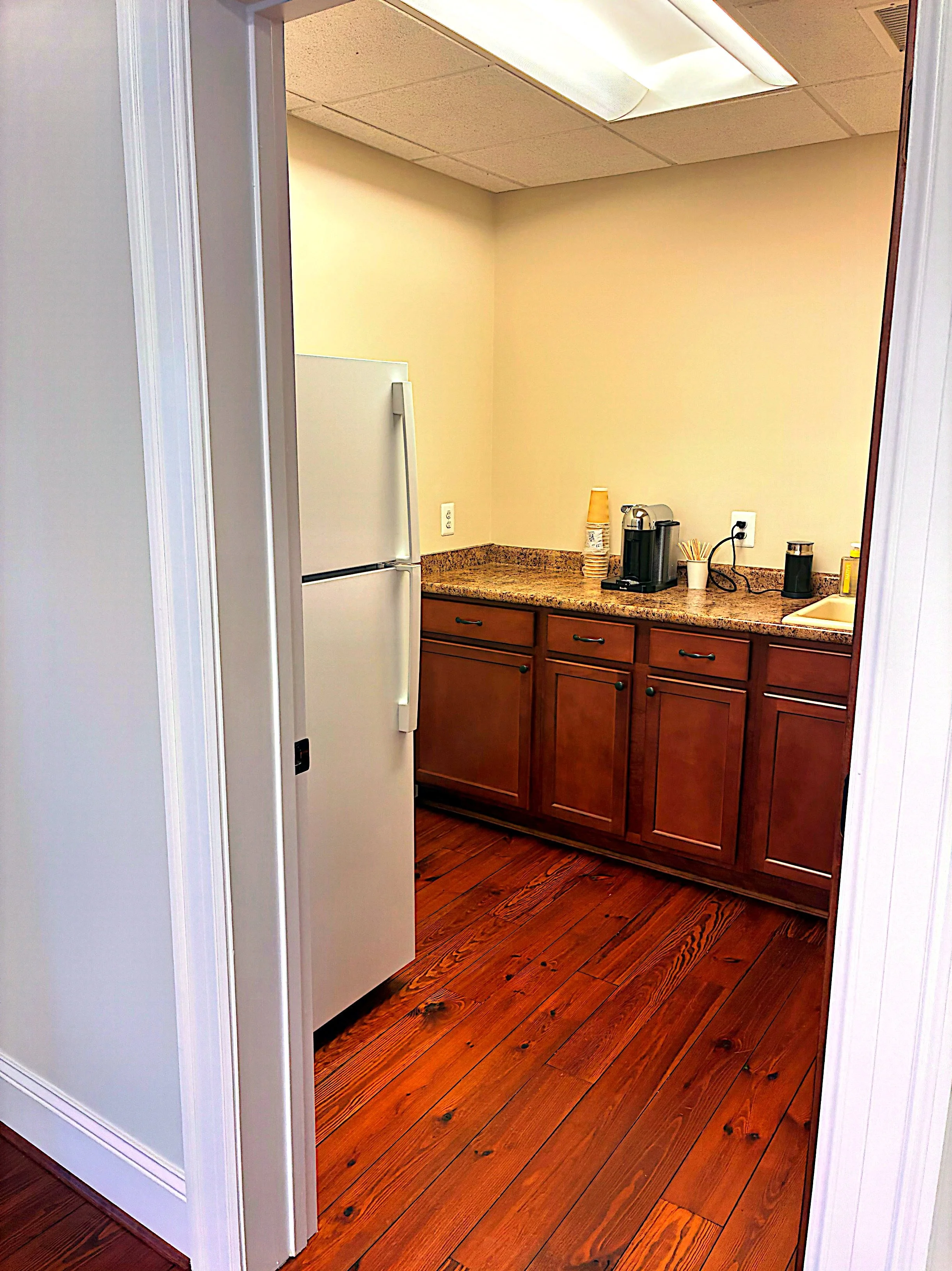 A kitchen with wooden cabinets, granite countertop, hardwood floor, and small appliances like a coffee maker and paper cups, seen through a doorway.