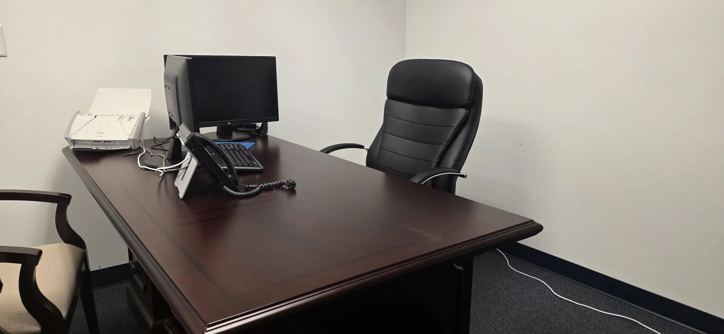 Empty office conference room with a wooden table, black office chair, a landline phone, computer monitor, keyboard, mouse, and printer.