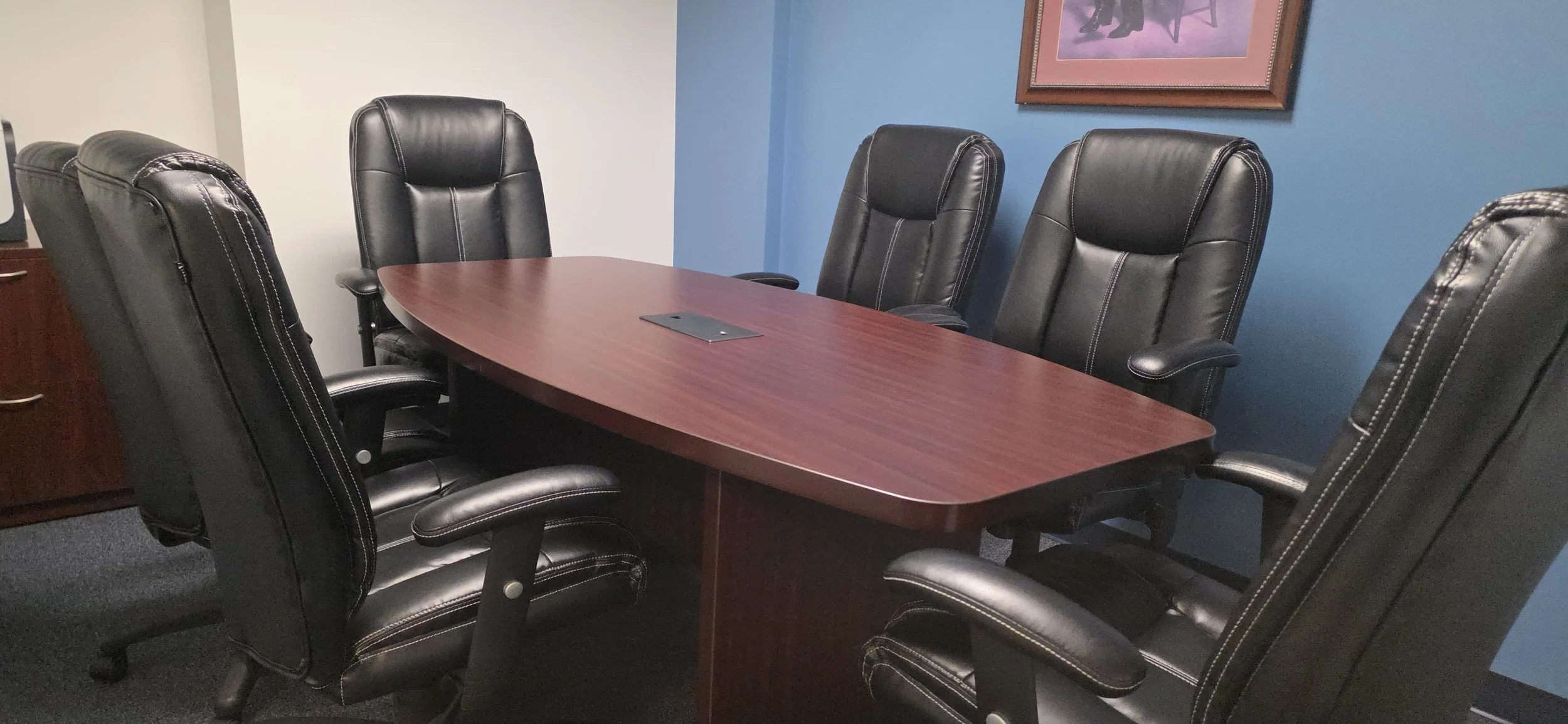A conference room with six black leather office chairs surrounding a wooden meeting table. The room has blue and white walls, and a framed picture with silhouettes of elephants hangs on the blue wall.