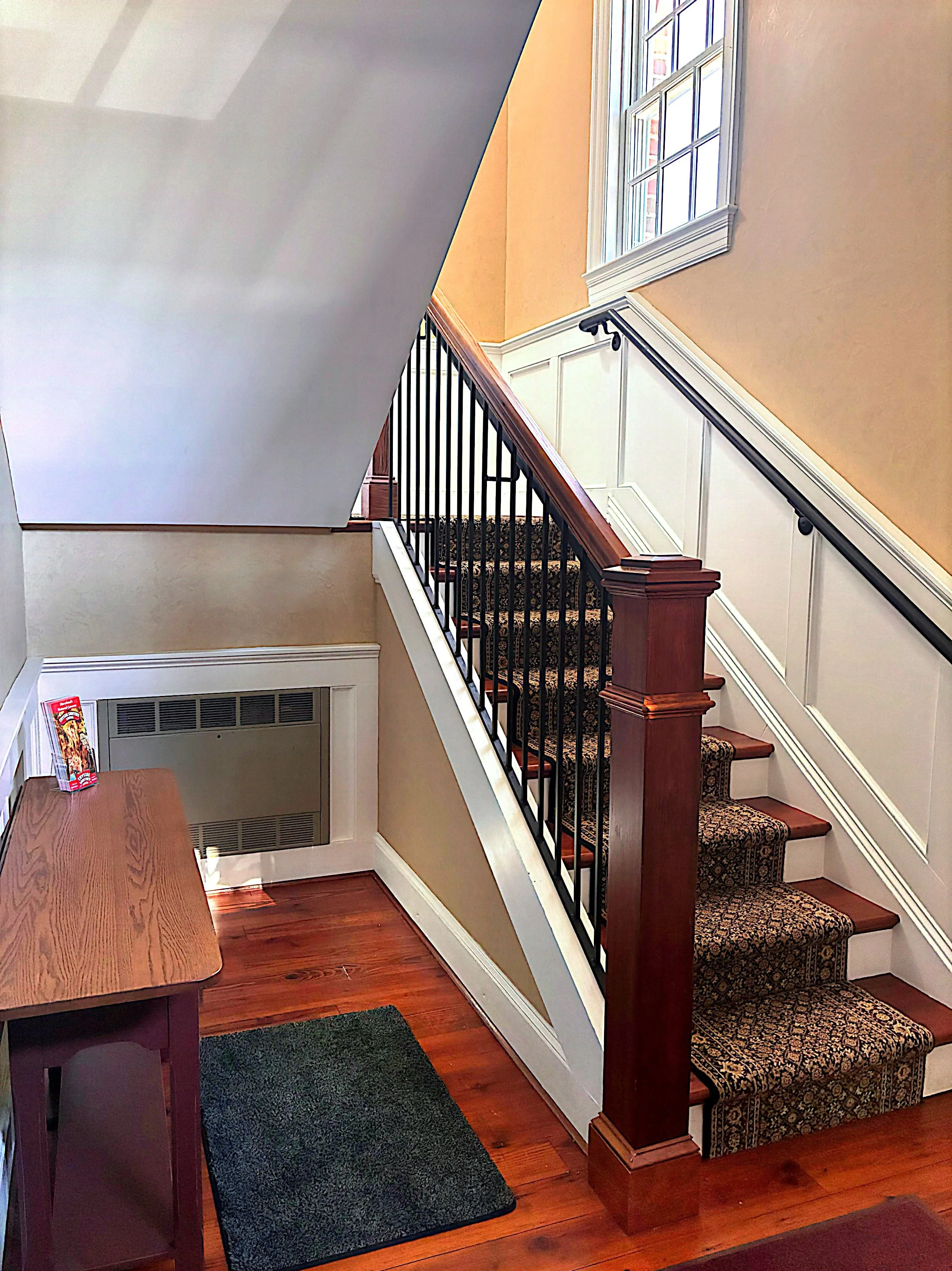 Interior view of a staircase with patterned carpeted steps, white paneling on the wall, wood handrail, and a large window. A wooden table with a brochure or flyer is visible on the left. The flooring is wooden, and there is a black rug at the bottom of the staircase.