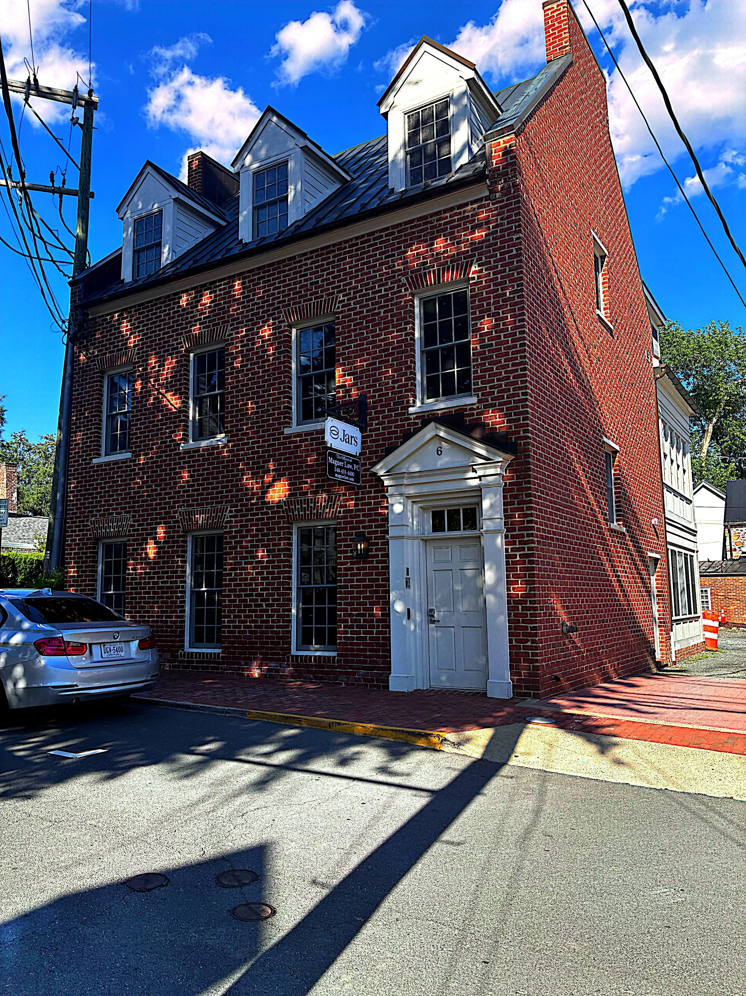 Red brick building with white roof and dormer windows, white door with decorative molding, parked silver car in foreground, blue sky with clouds, street with shadows and utility poles.