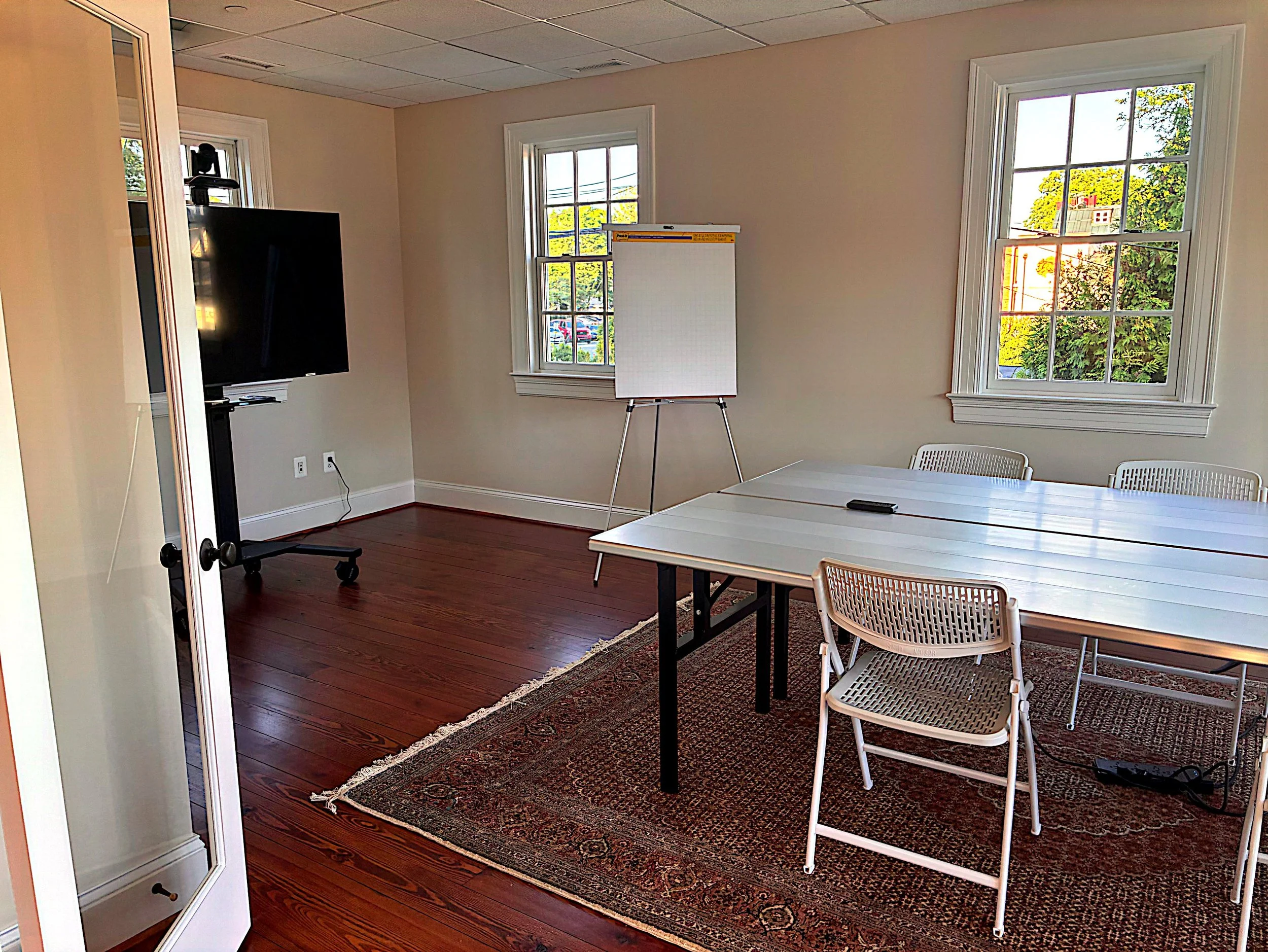 An office space with two large windows, a whiteboard on a stand, a table with chairs, a flat screen TV on a stand, and a patterned rug on wooden flooring.