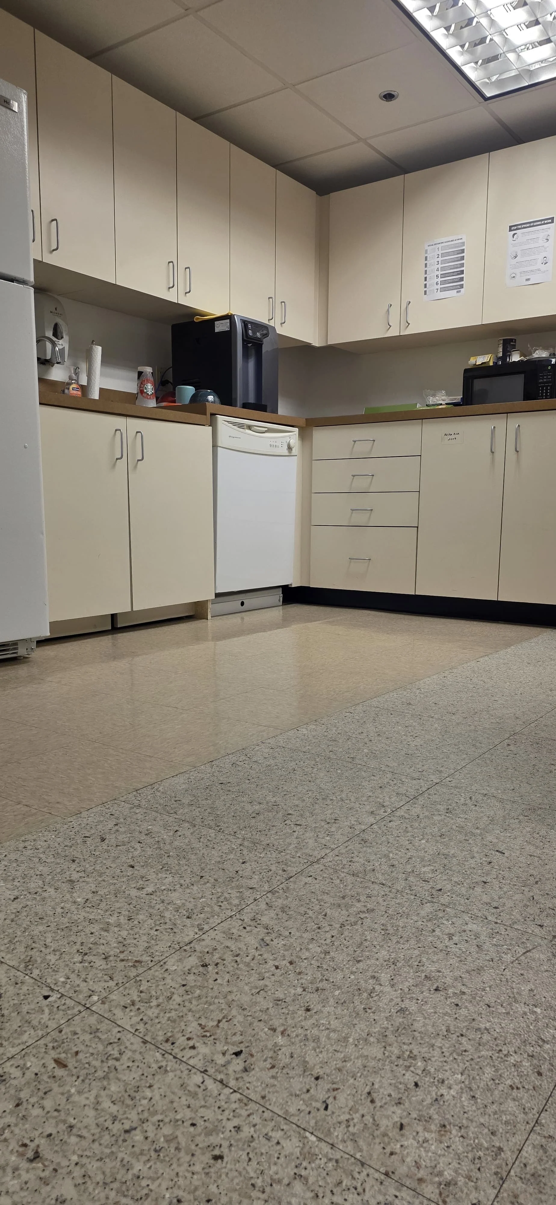 Office kitchen with beige cabinets, a dishwasher, microwave, coffee machine, and sink, with a linoleum tiled floor and ceiling lights.