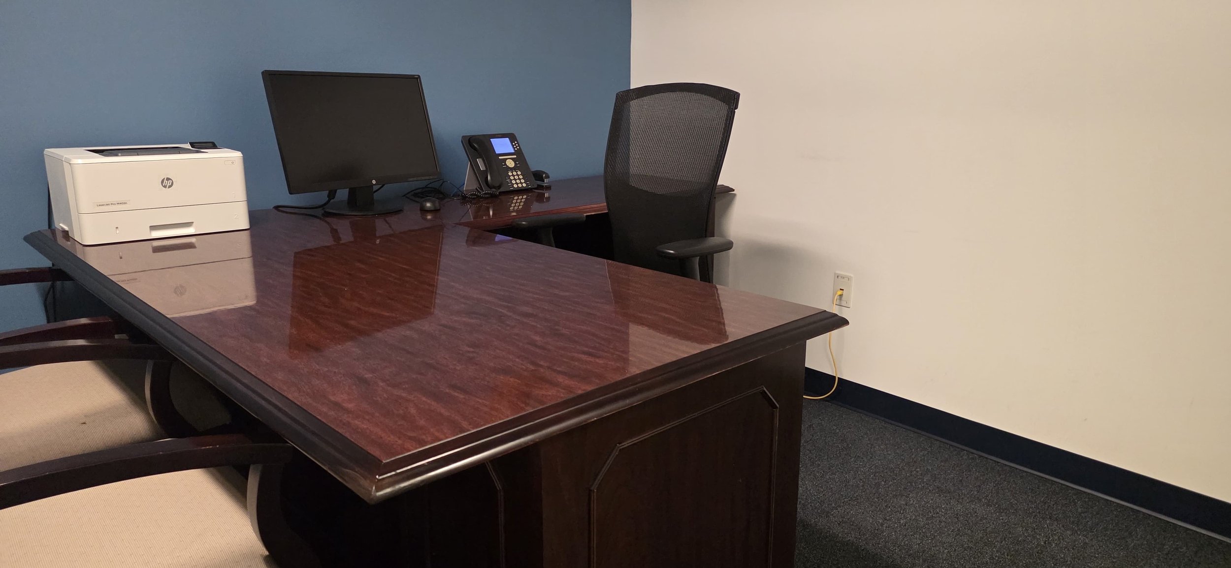 An office with a wooden desk, a computer monitor, a landline phone, a printer, and a gray office chair, with blue and white walls and a power outlet on the wall.