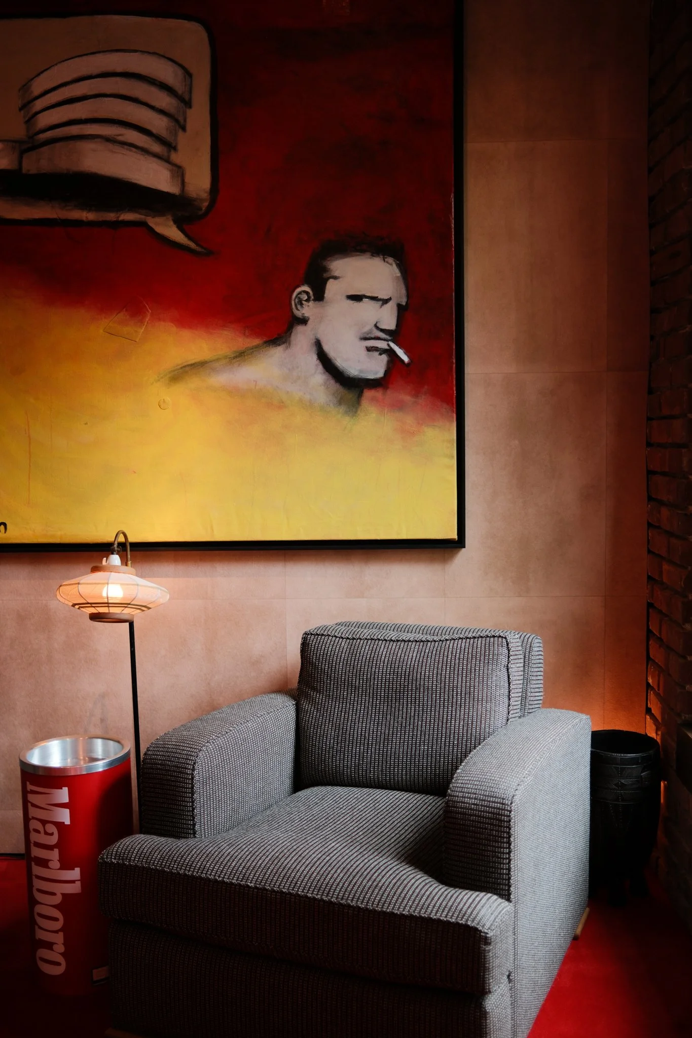 Blue armchair next to a floor lamp and Marlboro ash tray in front of a large red and yellow painting with man smoking cigarette