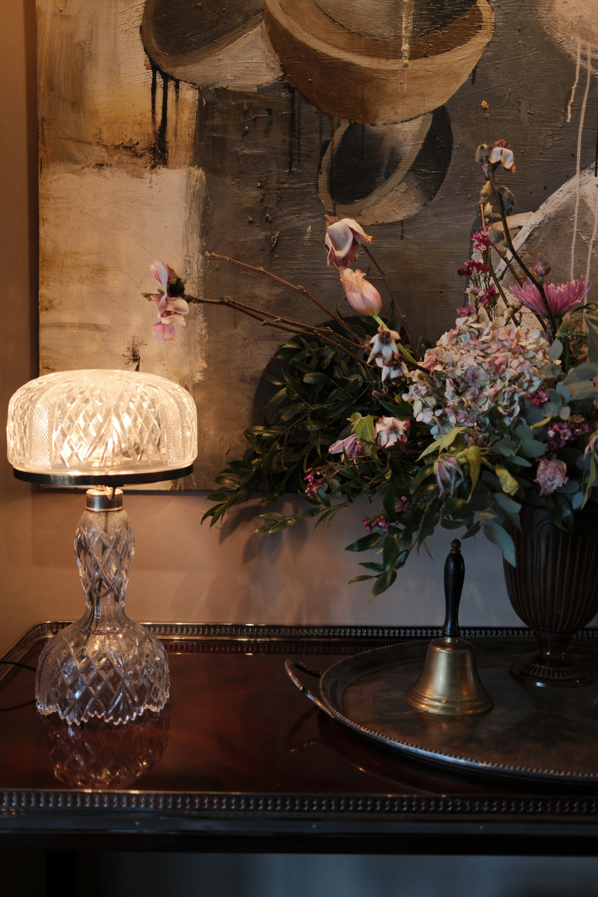 Art above a side table with a large floral arrangement, lamp, and bell