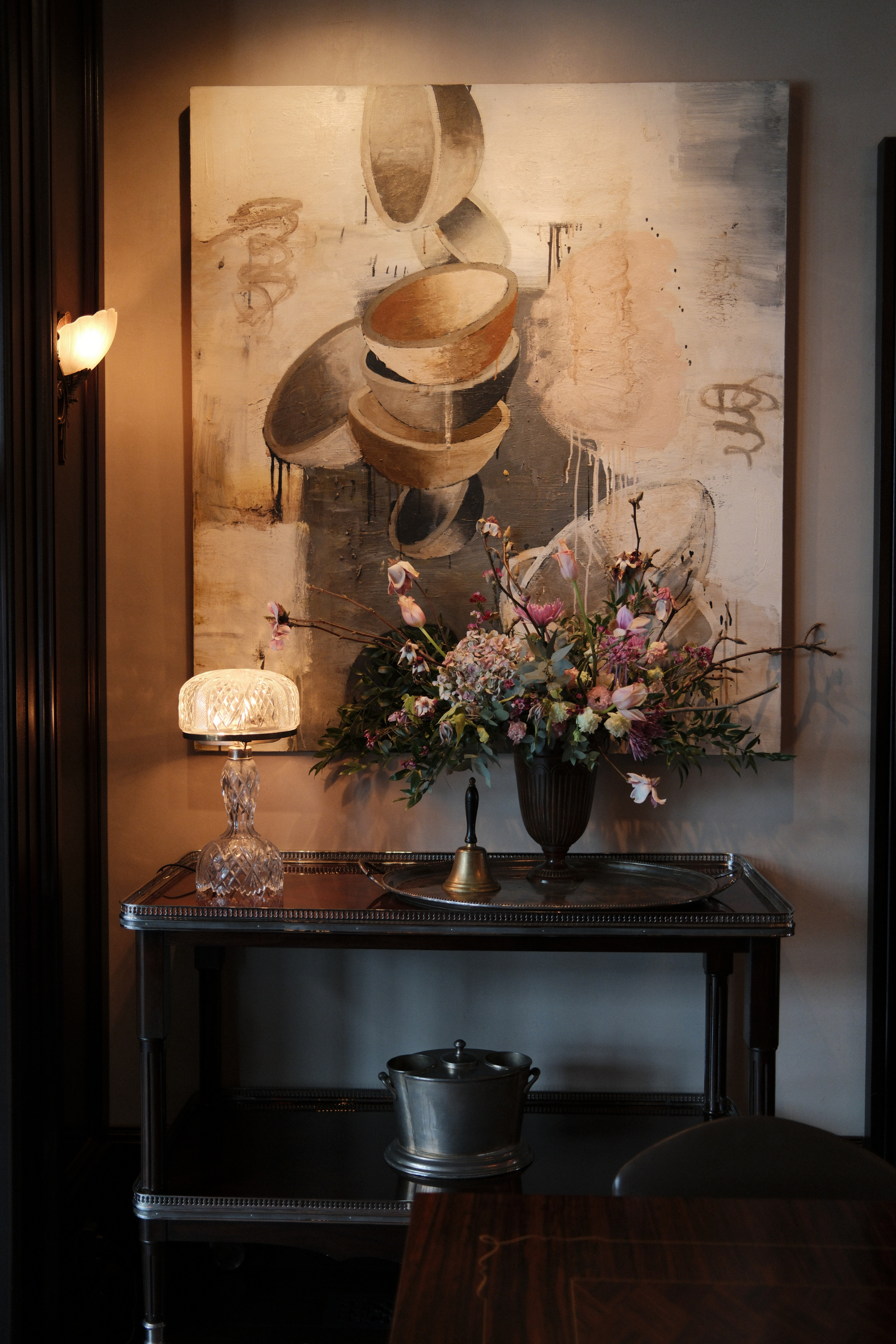 Art above a side table with a large floral arrangement, lamp, and bell