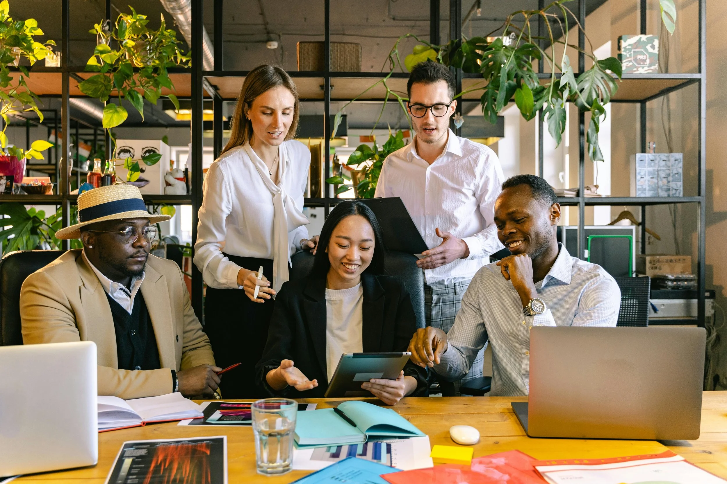 A group of five diverse coworkers working together in an modern office with green plants and shelves. Two women and three men are collaborating over a tablet and documents at a table with laptops and office supplies.