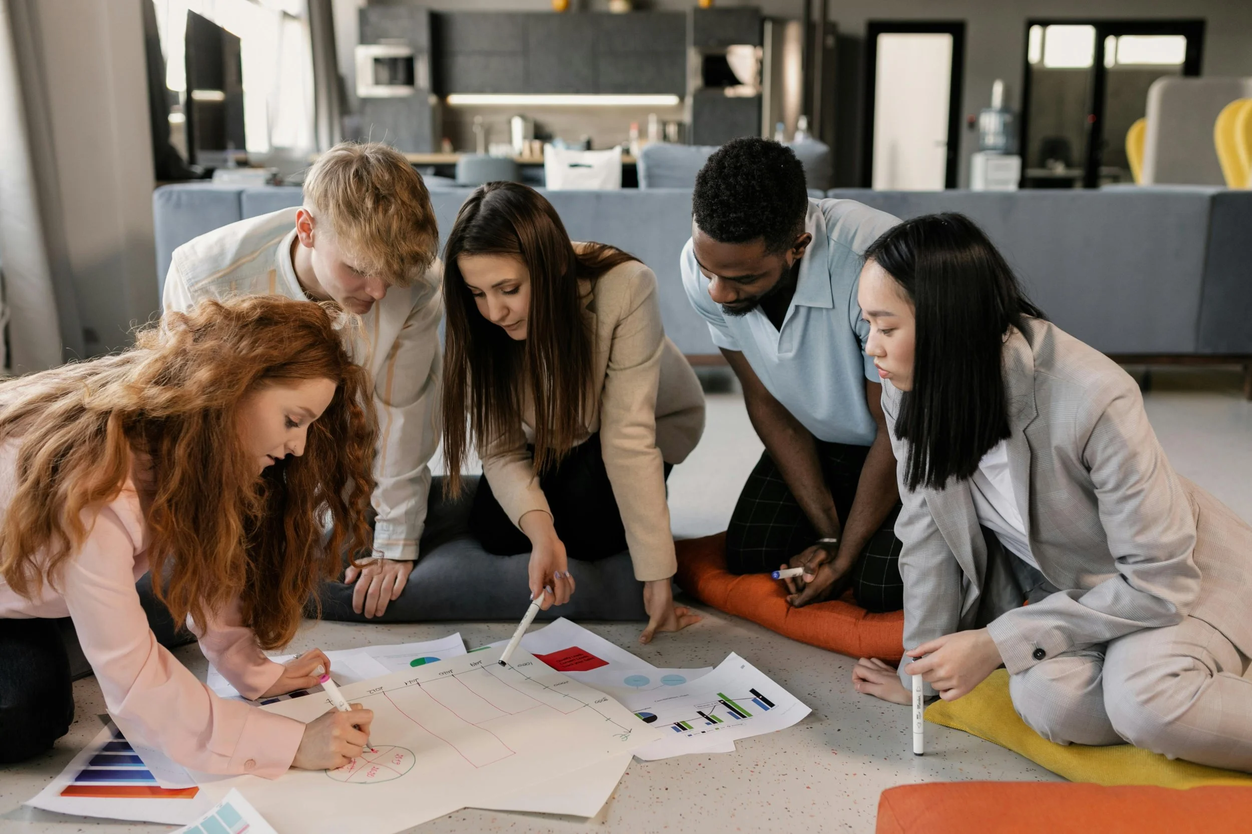 Group of six diverse young adults gathered around a table, working collaboratively on a project with charts, graphs, and markers in a modern office space.