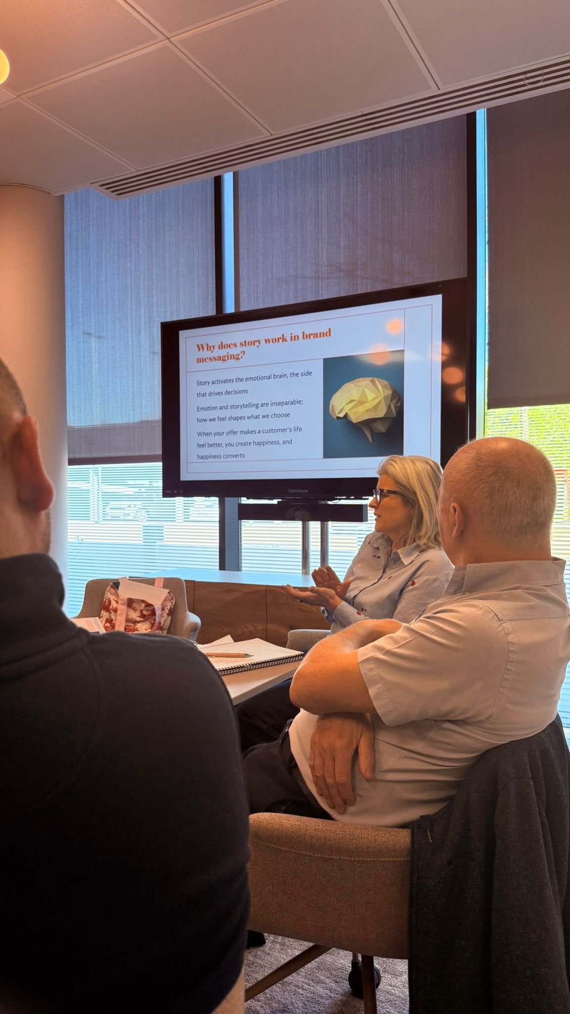 A group of people attending a presentation in a conference room, with a woman speaking and a large screen displaying a slide titled 'Why does story work in brand messaging?'.
