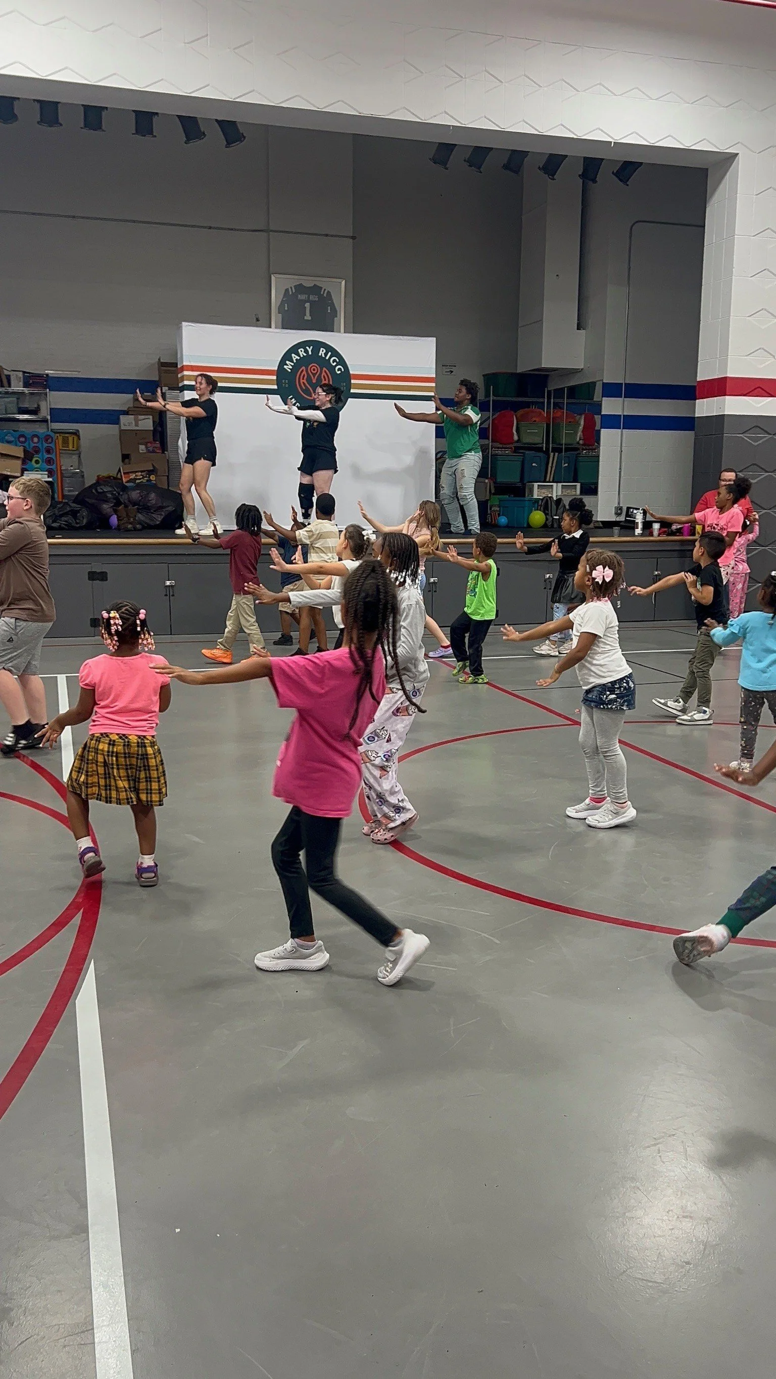 Children participating in a dance or exercise class in a gymnasium, led by instructors on a stage. The children are spread out and mirroring the instructors' movements.