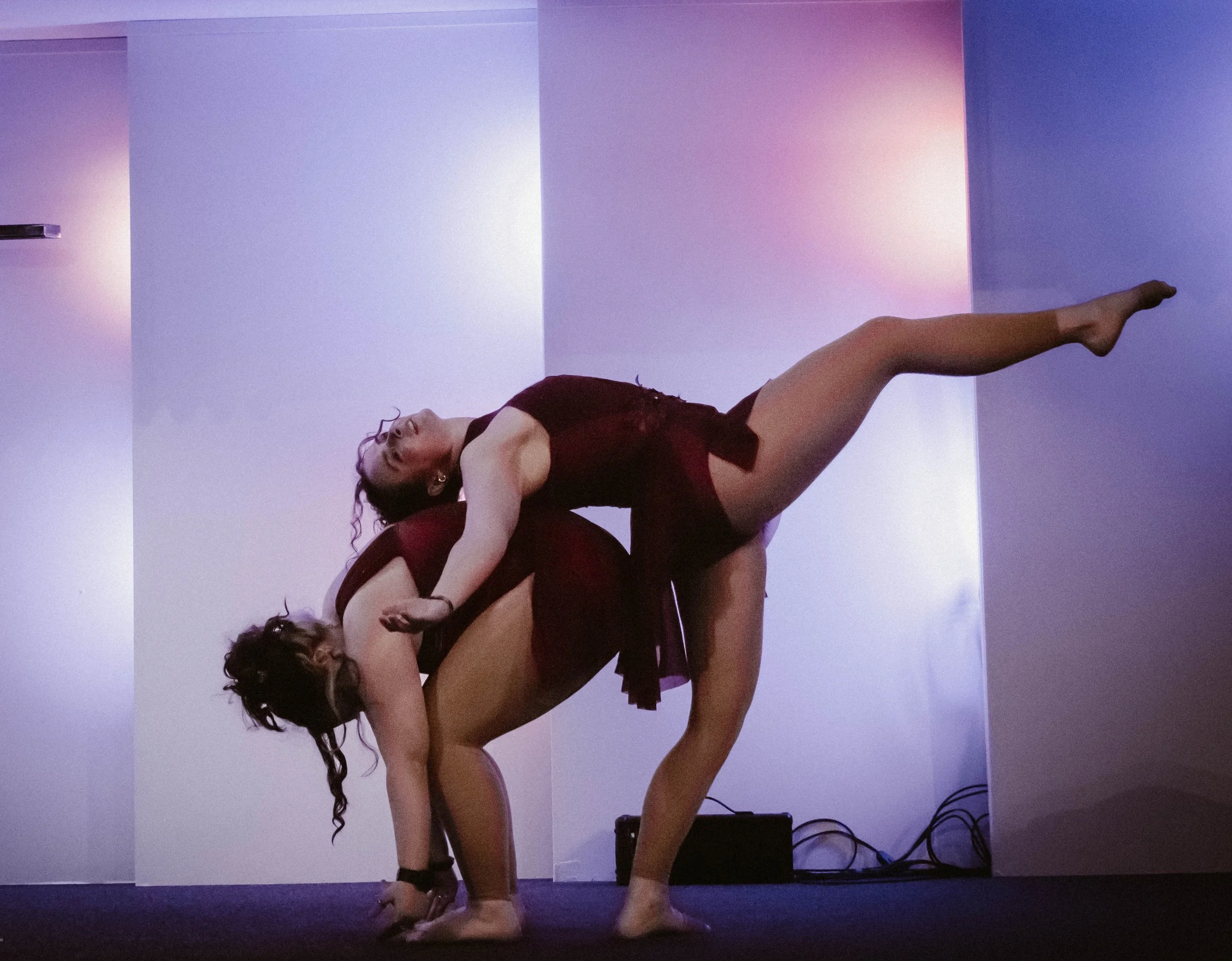 Two women wearing black dresses are performing a contortion pose in front of a wall with colorful lighting. One woman is standing on his hands while the other woman is leaning back across her body.