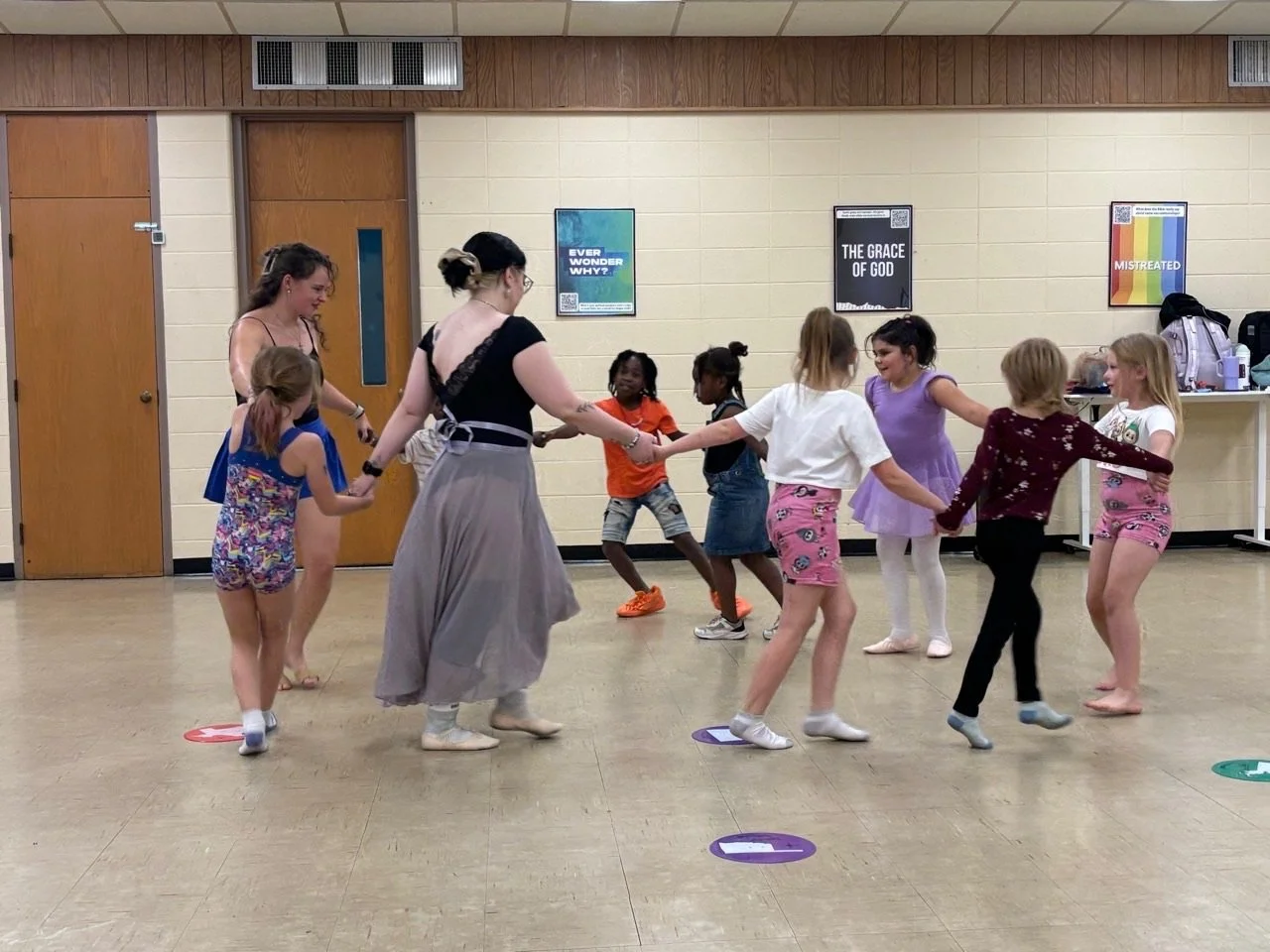 A group of children dancing and holding hands in a circle with two adults in a room with posters on the wall.
