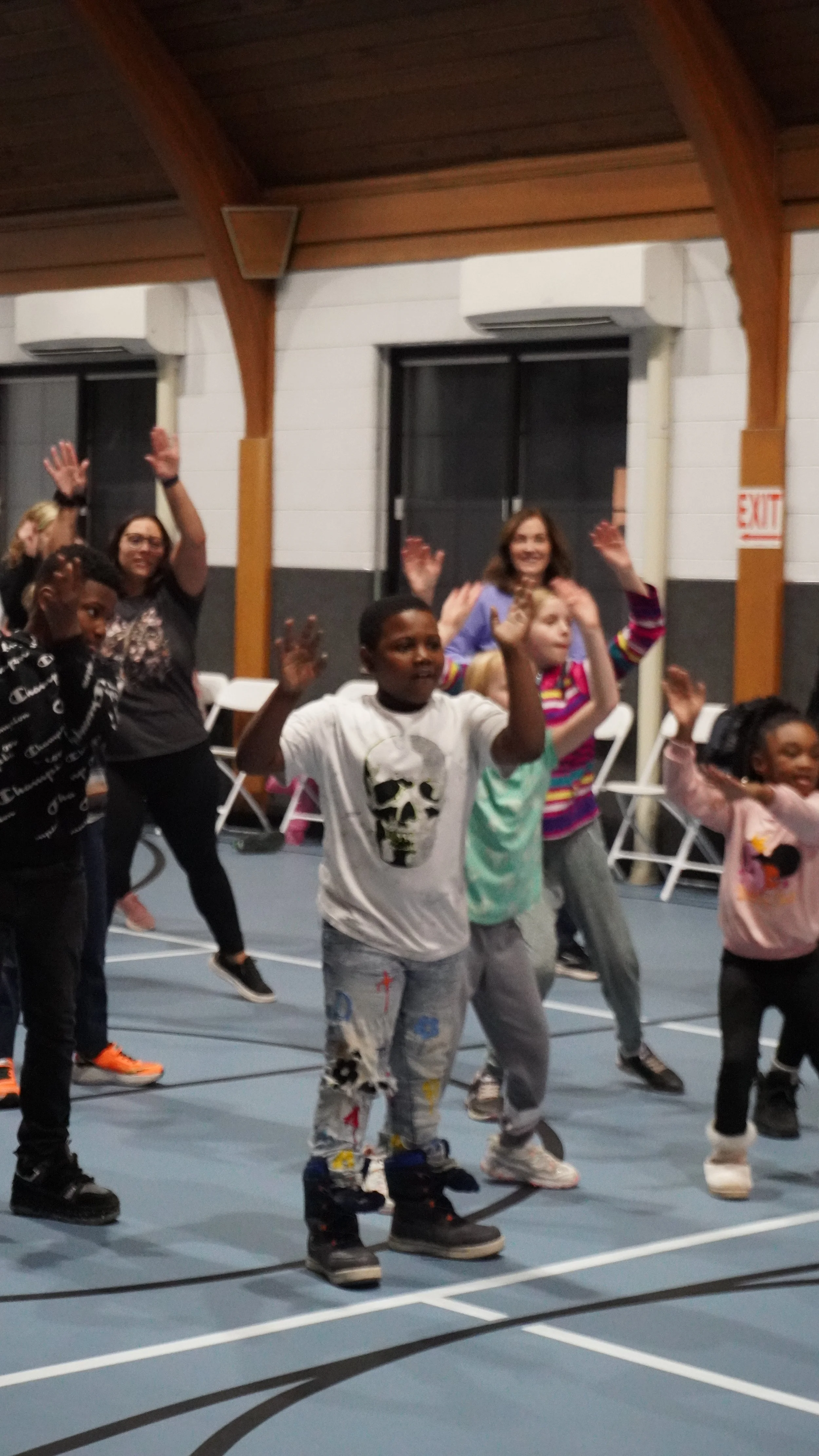 Children and adults dancing and enjoying a dance class in a gymnasium with wooden ceiling beams and white and black walls.