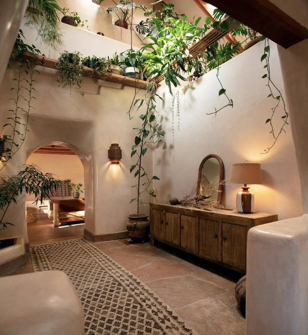 Cozy indoor space with white walls, wooden furniture, and lush hanging green plants. A mirror and table lamp are on a wooden sideboard, and a patterned rug runs along the floor. Warm lighting creates a welcoming atmosphere.