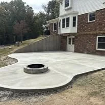 Concrete patio with a built-in circular fire pit in the center, adjacent to a brick and siding residential building, with a grassy yard and trees in the background.