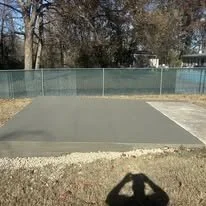 An empty tennis or basketball court with a chain-link fence and bare trees in the background.