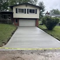 Newly poured concrete driveway in front of a two-story house with a garage, surrounded by grass and trees.
