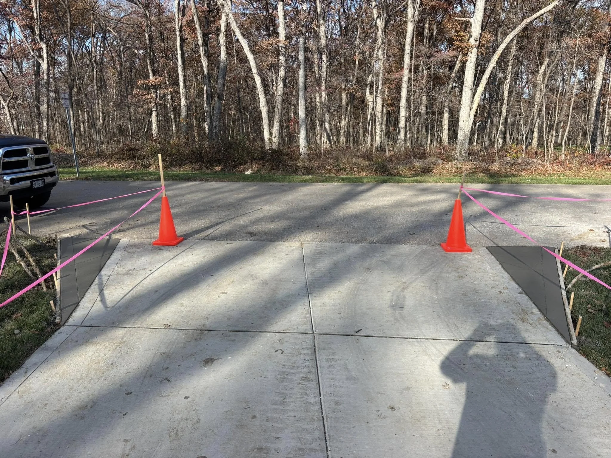 A newly constructed concrete driveway with orange traffic cones and pink caution tape, set beside a wooded area with leafless trees in the background.