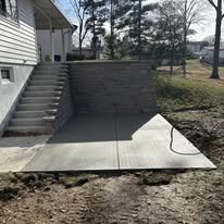 Backyard patio area with stairs, a brick wall, and a concrete slab, under a partly cloudy sky.