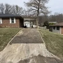 A sloped driveway with concrete and brick sections leading up to a house, with grass on either side and trees in the background.