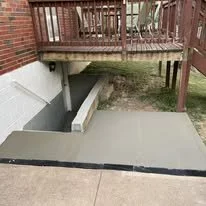 Concrete pathway leading to a backyard with a wooden deck above. The area below has a pipe and a small grassy patch.