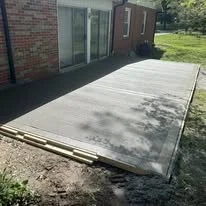 A plastic outdoor patio or deck cover installed over a concrete slab next to a brick house with a sliding glass door, in a yard with grass and trees.