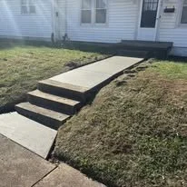 Concrete ramp with stairs leading up to a house, surrounded by grass.
