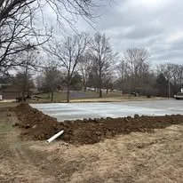 A construction site with a freshly poured concrete slab and surrounding dirt, in a park-like area with leafless trees.