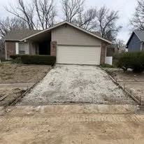 A newly poured concrete driveway in front of a house with a white garage door, surrounded by a yard with some bushes and trees.