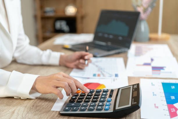 Person using a calculator at a desk with financial documents, a laptop, and a pen.